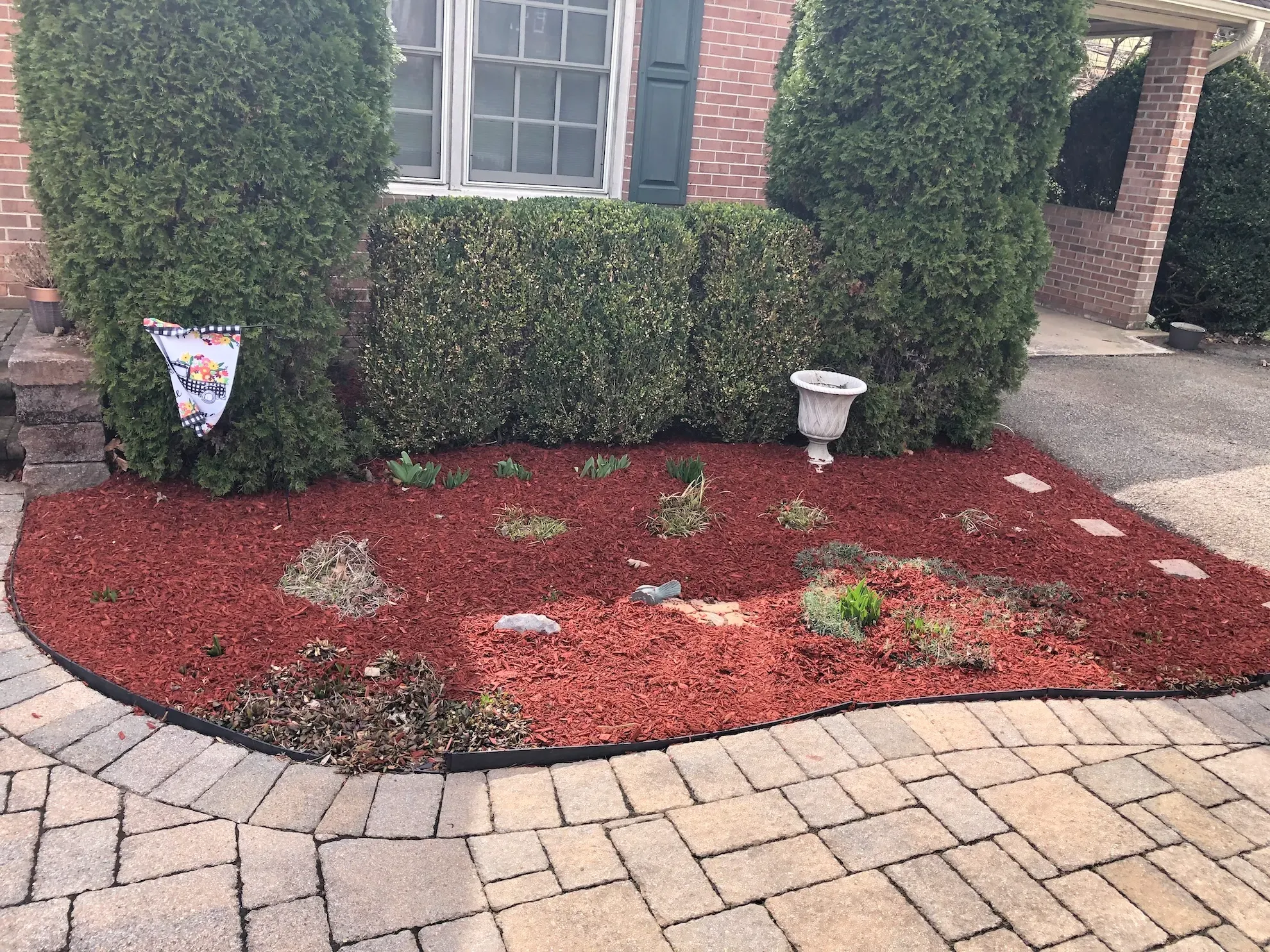A garden with red mulch and a vase in front of a brick house.