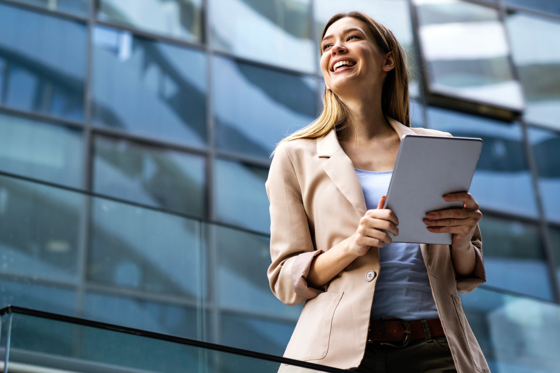 Woman in business attire, holding tablet, smiles in front of glass building.