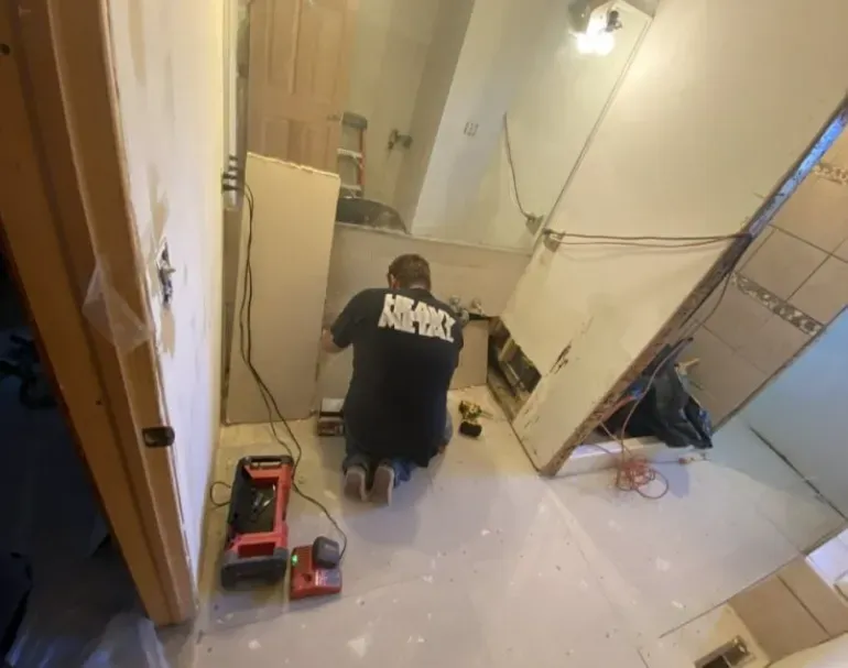 Person kneels, cutting drywall in a bathroom under renovation. Tools and construction materials are scattered around.