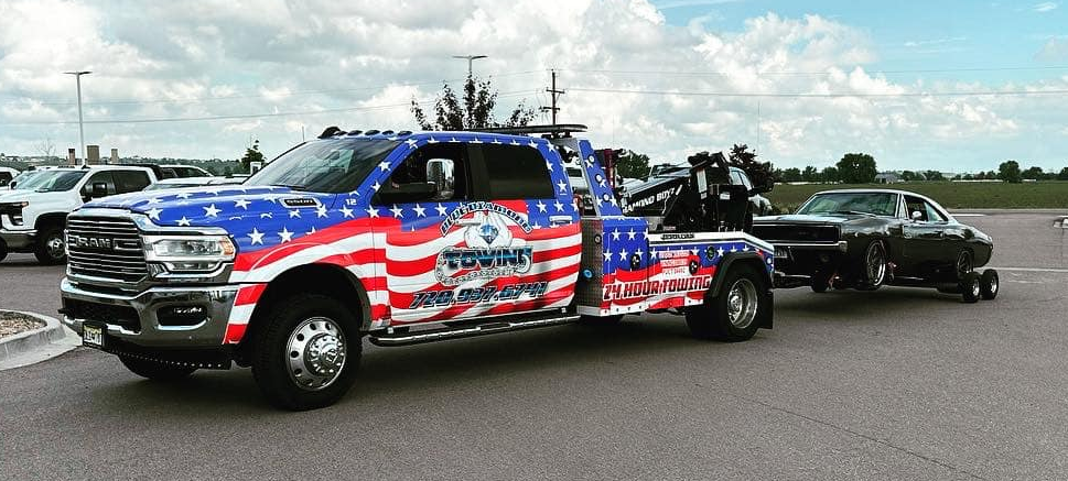 Tow truck, American flag design, towing a black car on a paved lot.