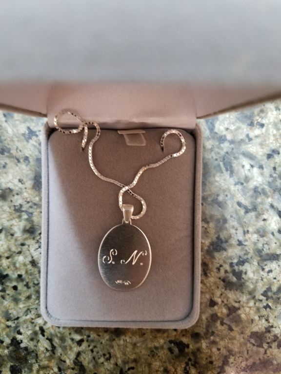 Oval silver necklace pendant with engraved initials in a small open box on a speckled countertop