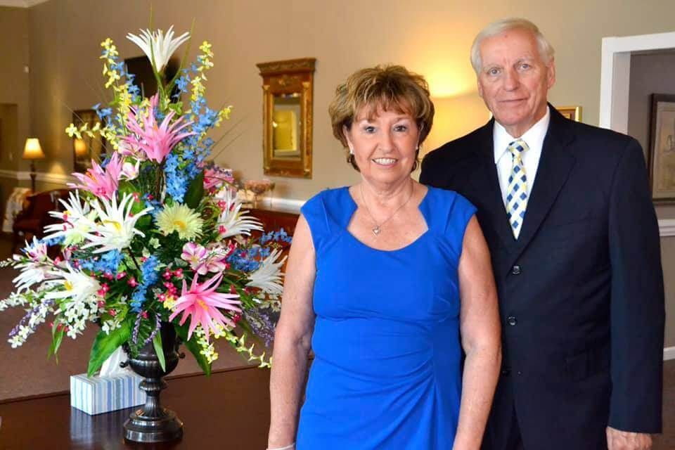 Two people standing indoors beside a large colorful flower arrangement and a mirror.