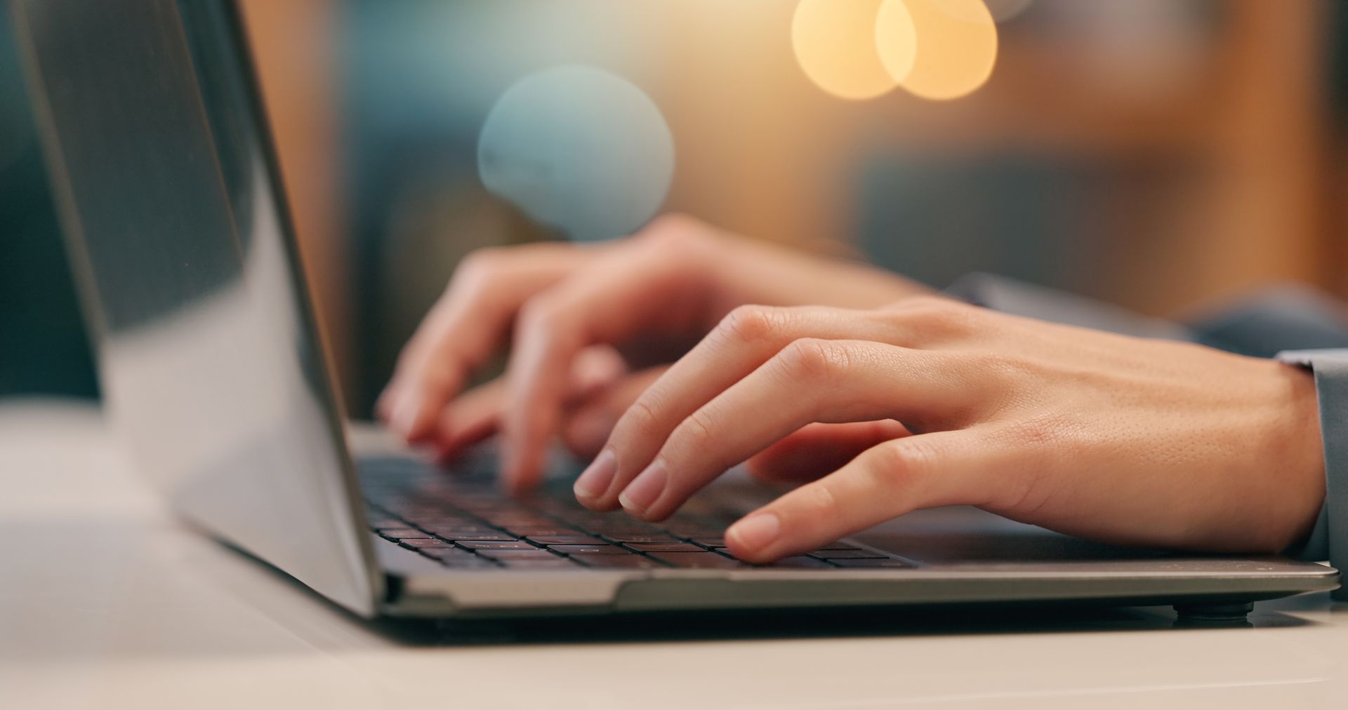 Hands typing on a laptop keyboard beside a blurred coffee cup in warm indoor light