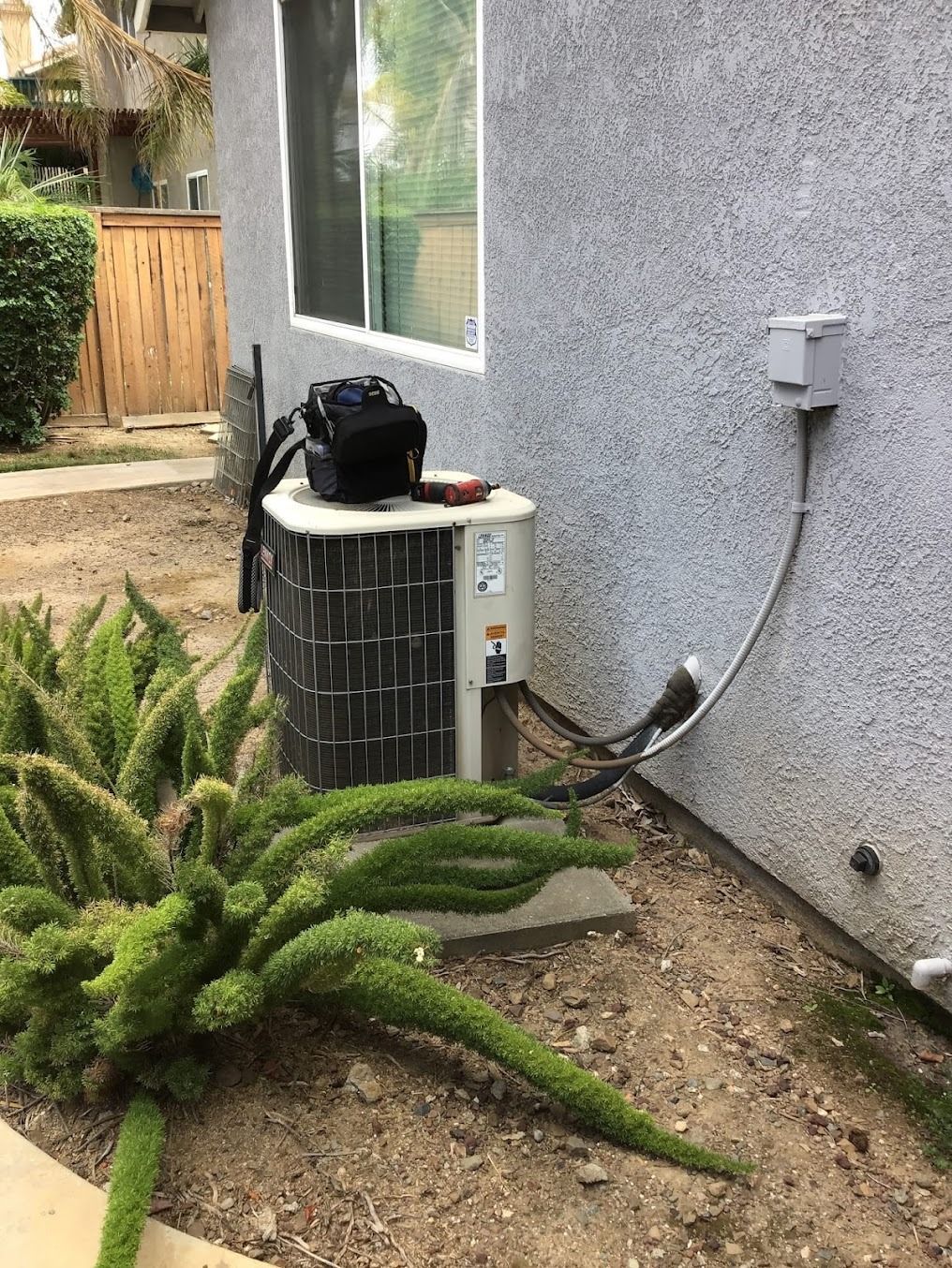 Outdoor air-conditioning unit beside a gray house wall, with hose and plants in the foreground.