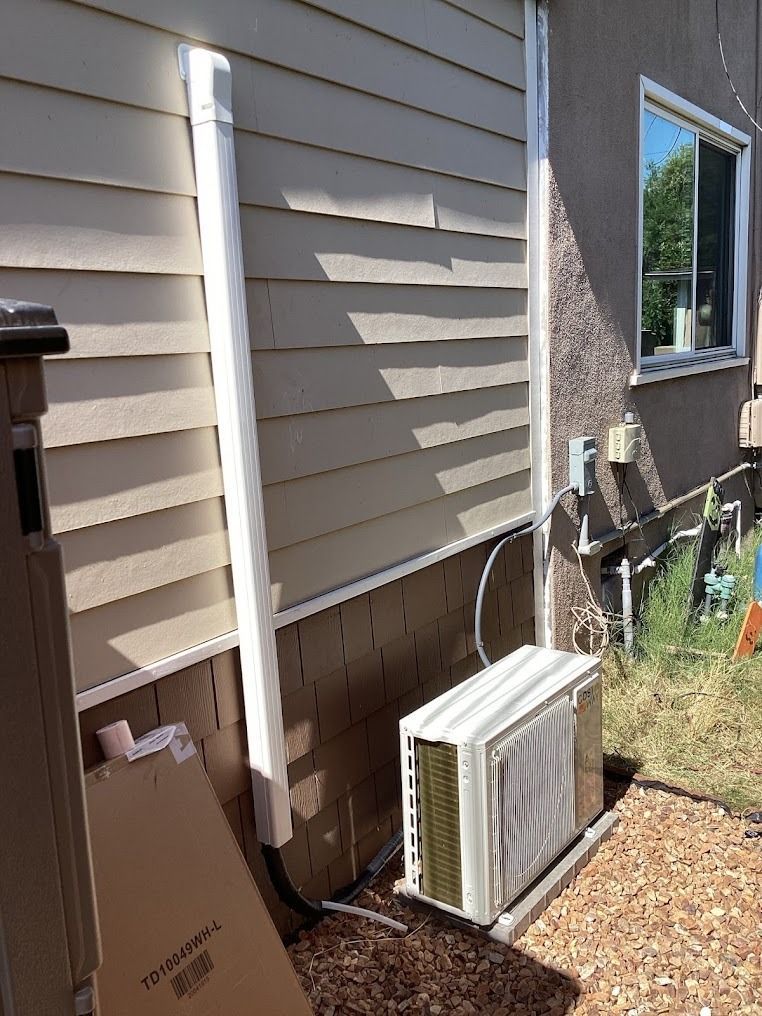 Side of a house with white siding, a window AC unit on the ground, and a white downspout by the foundation.