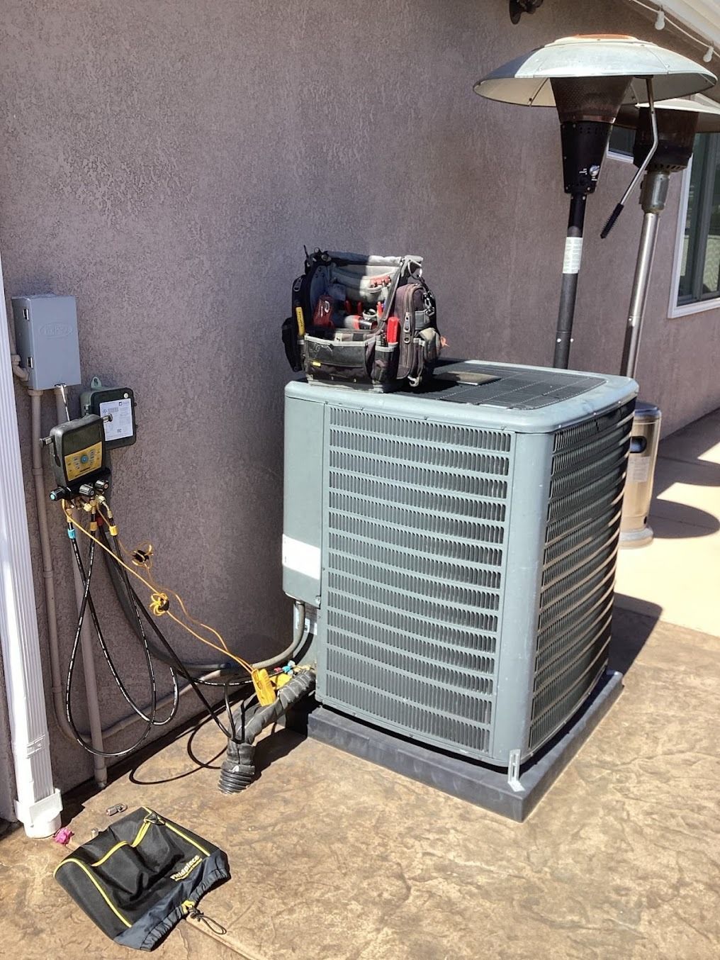 Outdoor HVAC unit with service tools and cables beside a house wall and patio heater