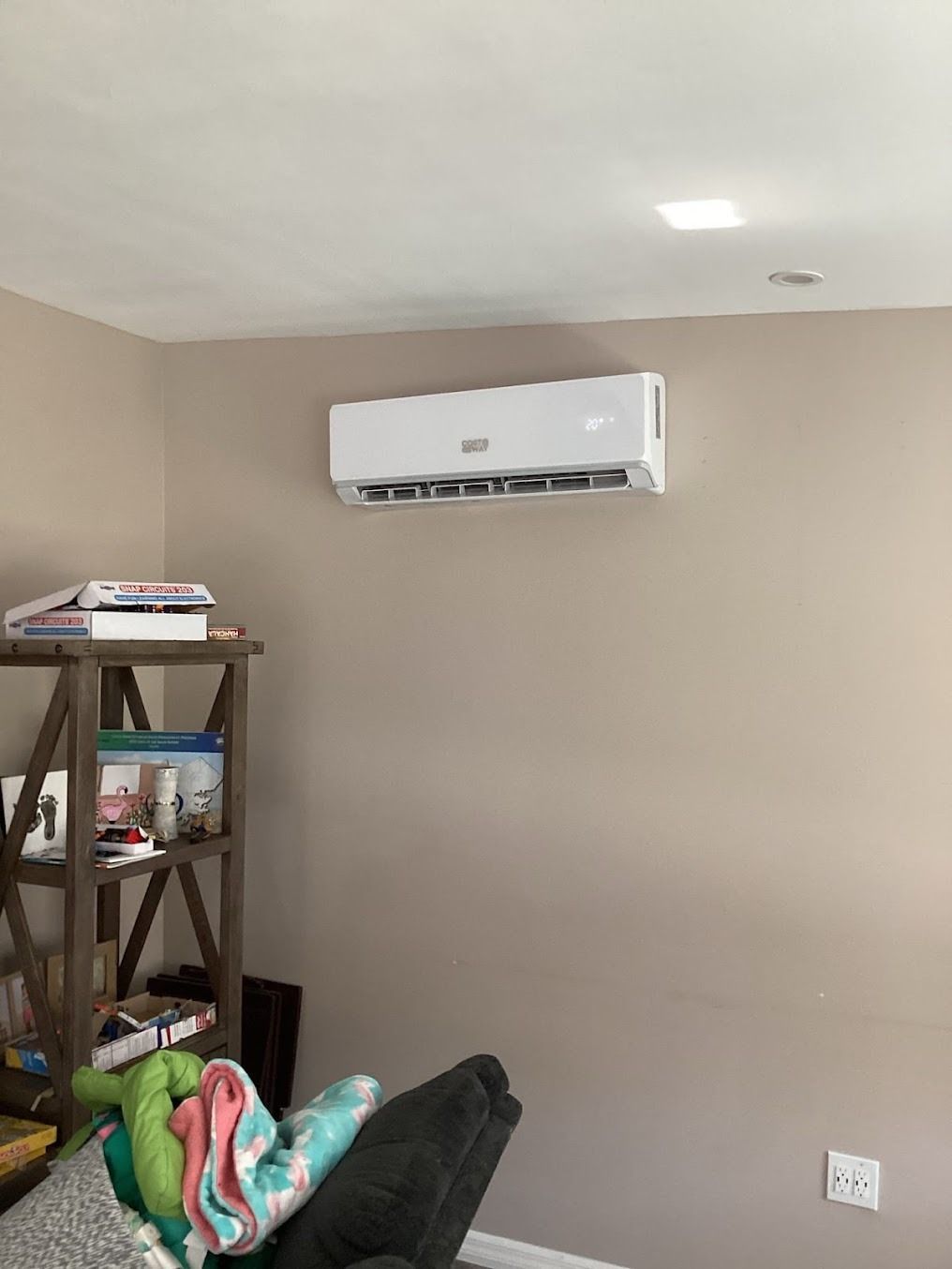 Small beige room with a wall-mounted white air conditioner and a shelf with folded items on the left.