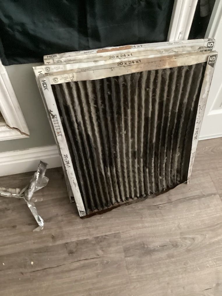Dirty HVAC air filter leaning on a hardwood floor, with a small detached metal piece nearby