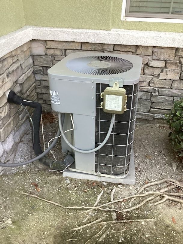 Outdoor air conditioner unit beside a stone wall with exposed roots in the yard