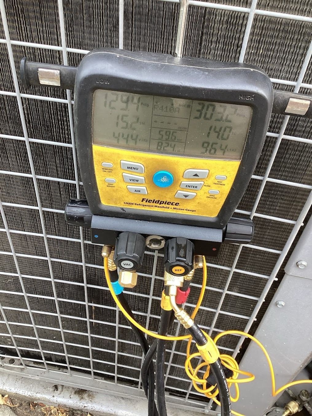 Digital HVAC gauge with yellow control panel and attached hoses on an outdoor unit grille.