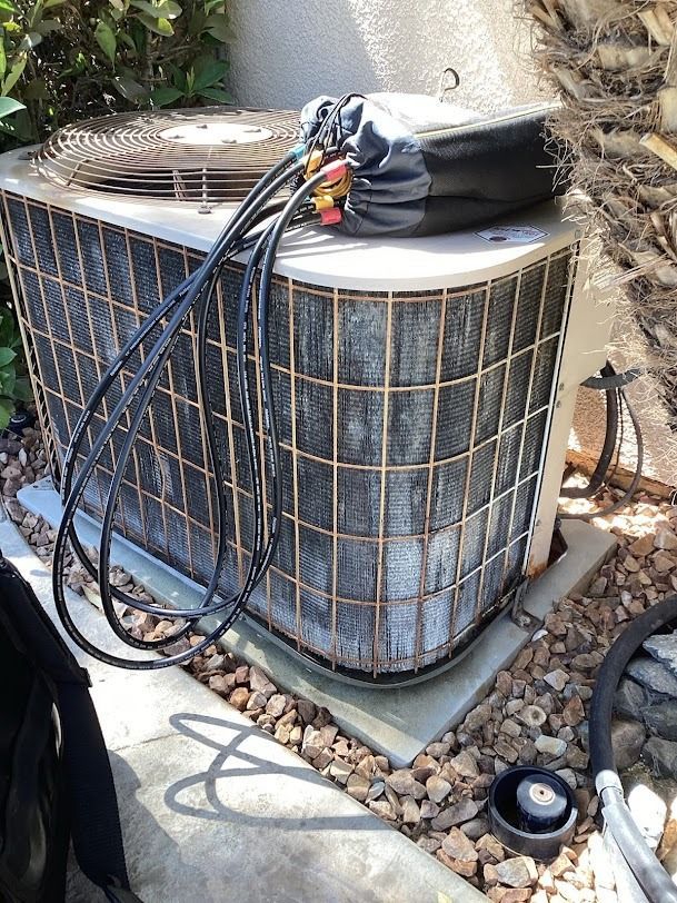 Outdoor HVAC unit with hoses and a tool bag on top, beside a wall and gravel ground