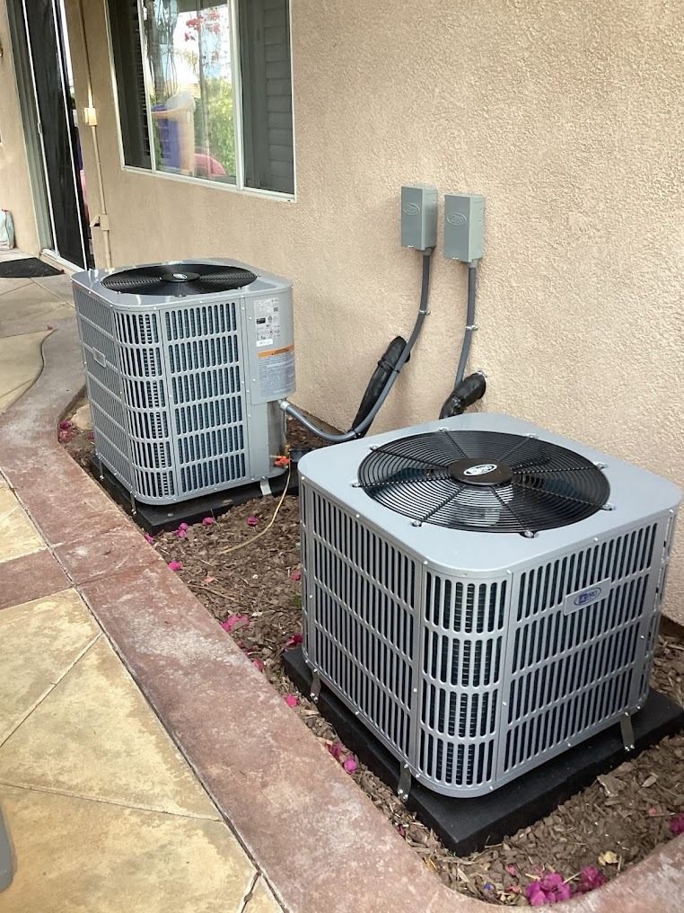 Two outdoor HVAC condenser units beside a beige house wall on a paved side yard.