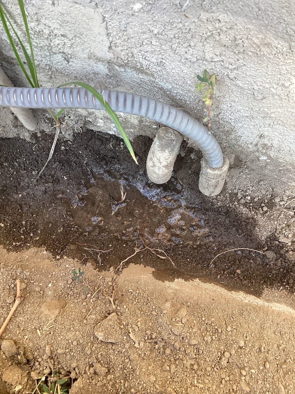 Gray corrugated hose beside a wall, above a dark soil trench and dry dirt ground