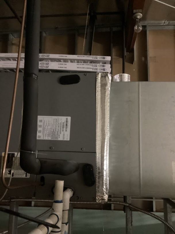 Gray HVAC unit and ductwork mounted on an unfinished ceiling with exposed pipes and wires