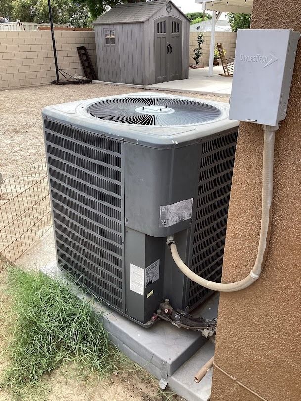 Outdoor air conditioner unit beside a tan house wall, with a gray shed and patio in the background