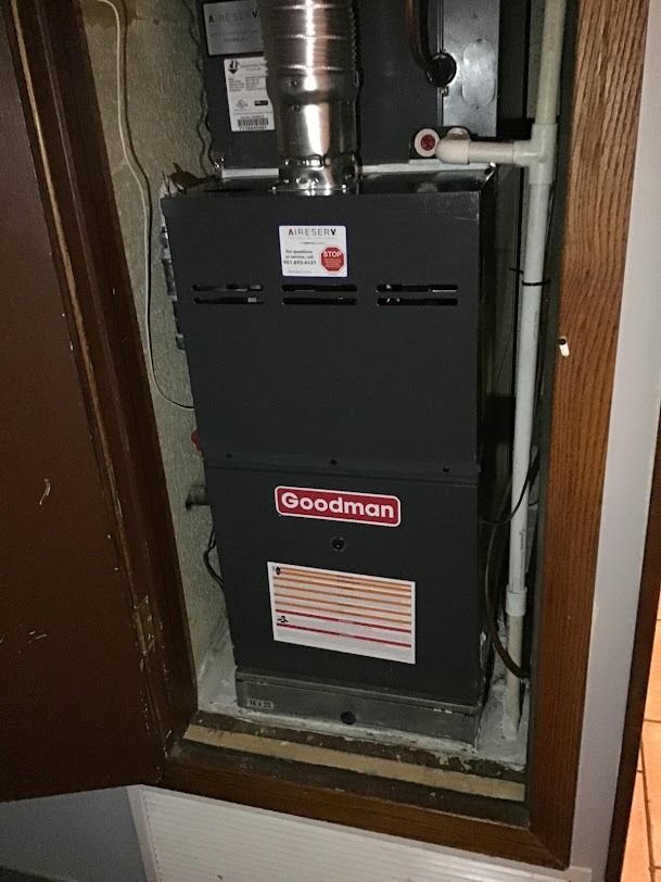 Black Goodman furnace installed in a narrow utility closet with exposed ductwork and pipes.