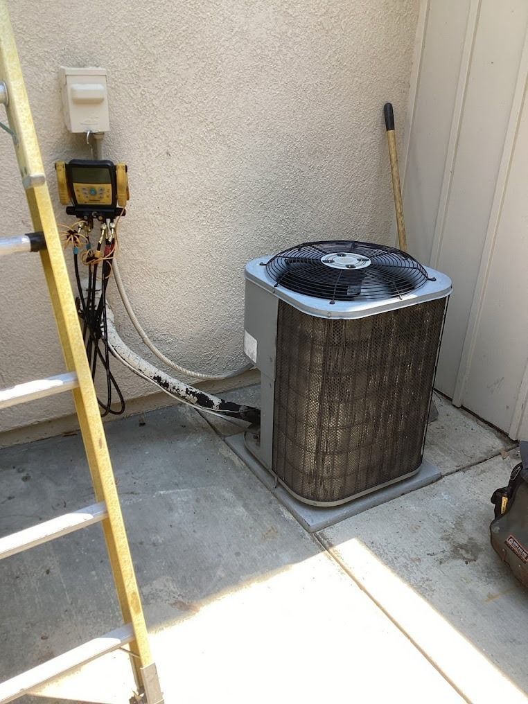 Outdoor HVAC condenser unit beside a stucco wall, with tools and a ladder nearby.