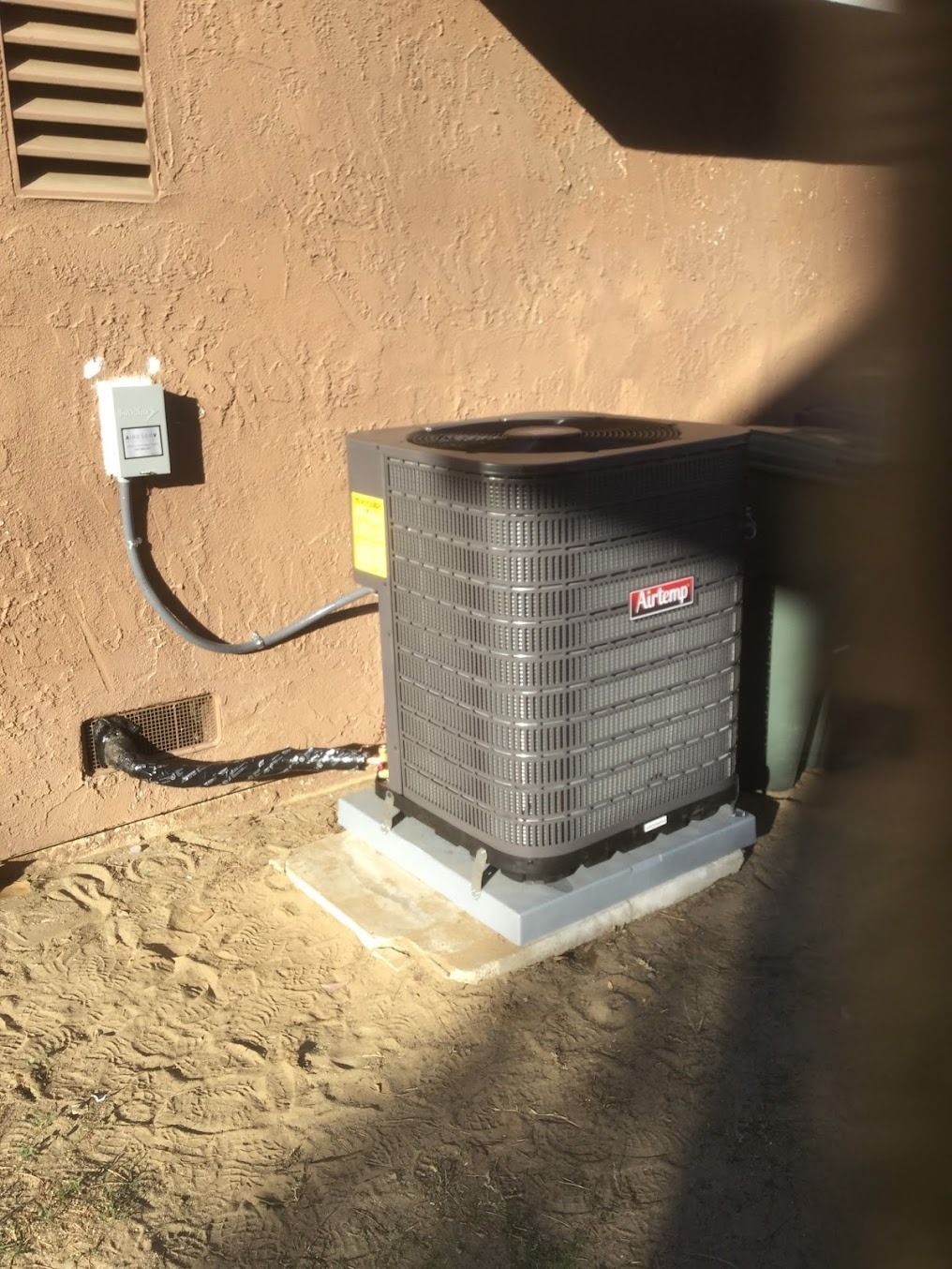 Outdoor air conditioner unit beside a stucco wall and electrical box, partly in shade.