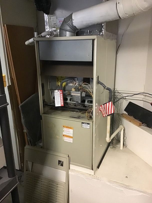Large gray HVAC furnace in a utility room with exposed ducts and pipes.