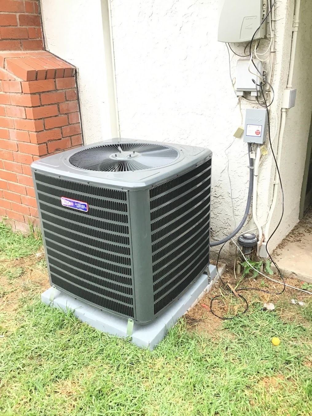 Outdoor air conditioner unit beside a brick wall and electrical meter, on grass.