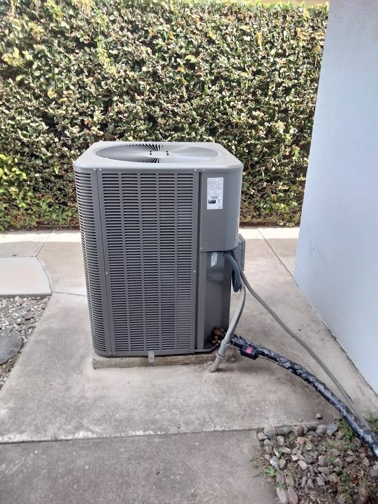 Outdoor air conditioner unit beside a wall on a concrete patio, with hedge in the background.