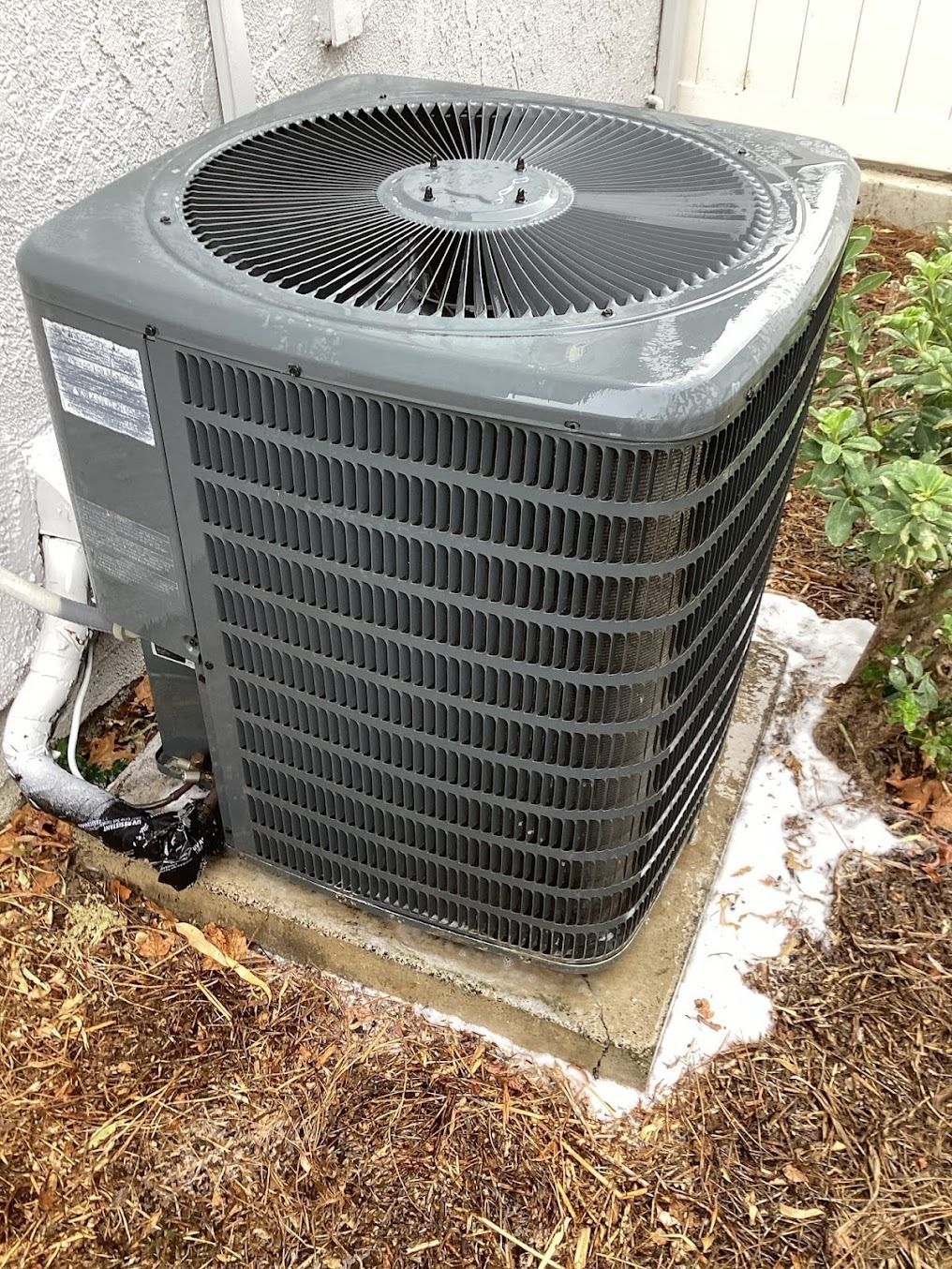 Outdoor air conditioner unit sitting on a concrete pad beside mulch and grass, with light debris on top.