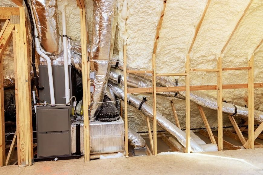 Insulated attic with spray foam, ductwork, and HVAC equipment on a wooden framing grid