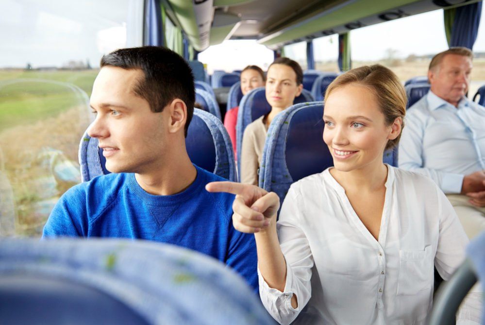 People on a bus, a woman points to something outside the window while looking at a man beside her.