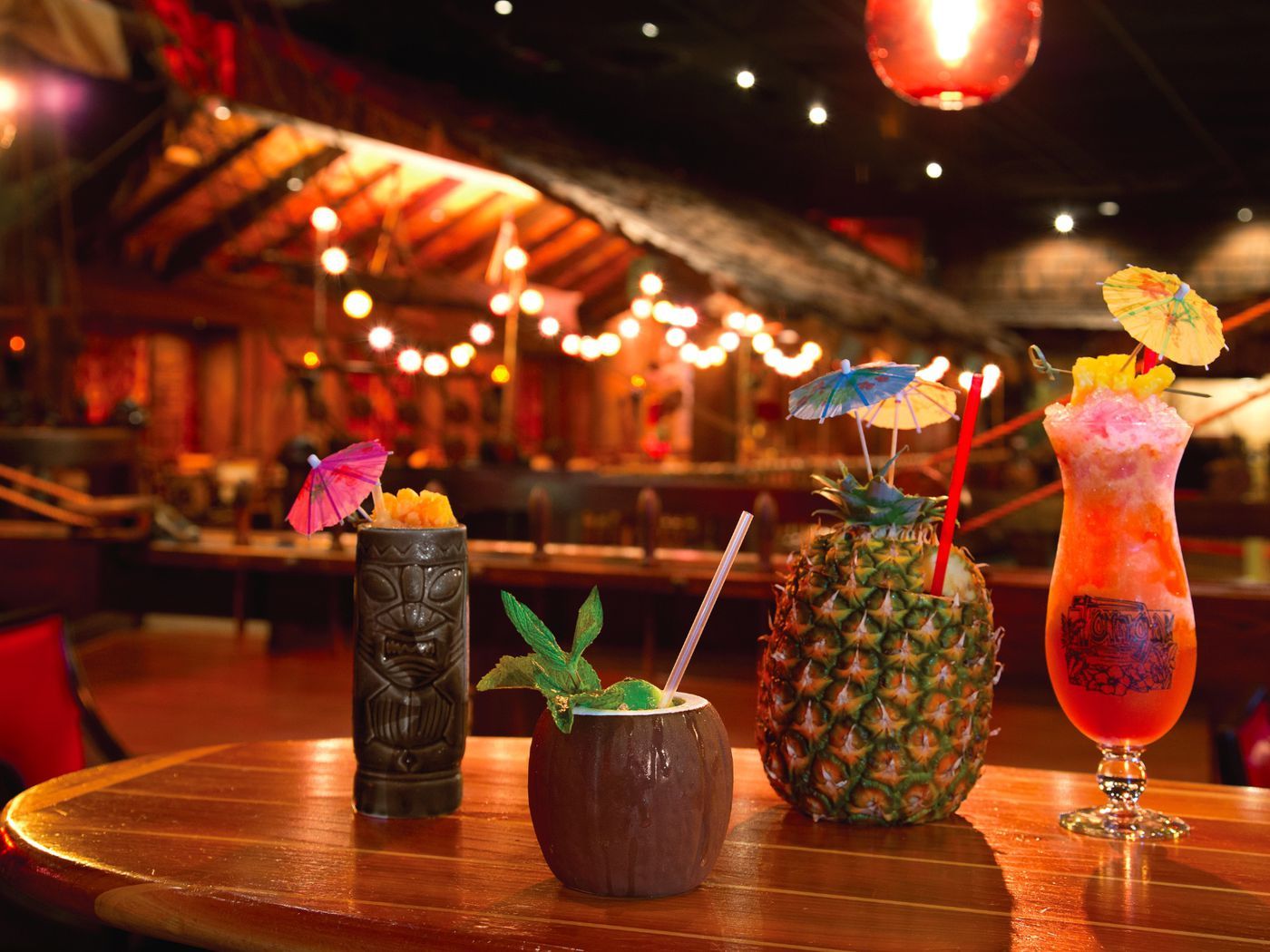 Four tiki cocktails on a wooden table in a dimly lit tropical bar.