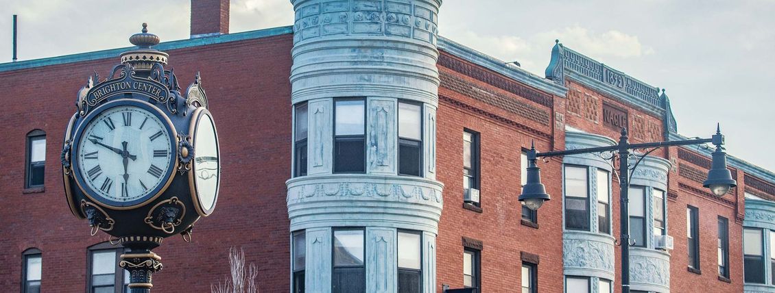 Ornate clock in front of a brick building with decorative details and a street lamp.