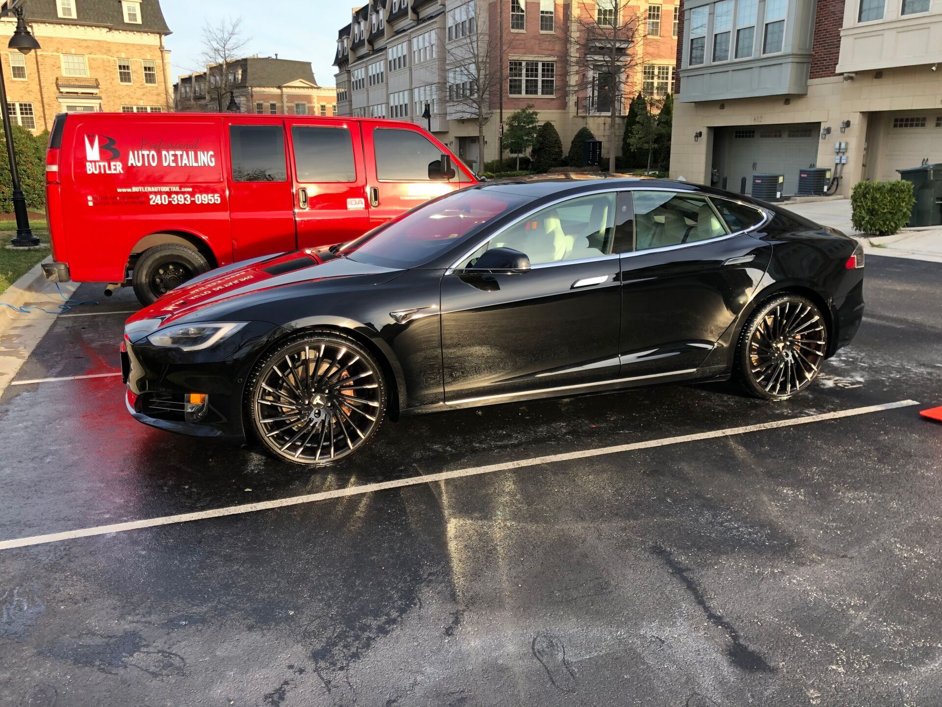 Black Tesla sedan parked, next to a red detailing van, in a paved lot with wet pavement.