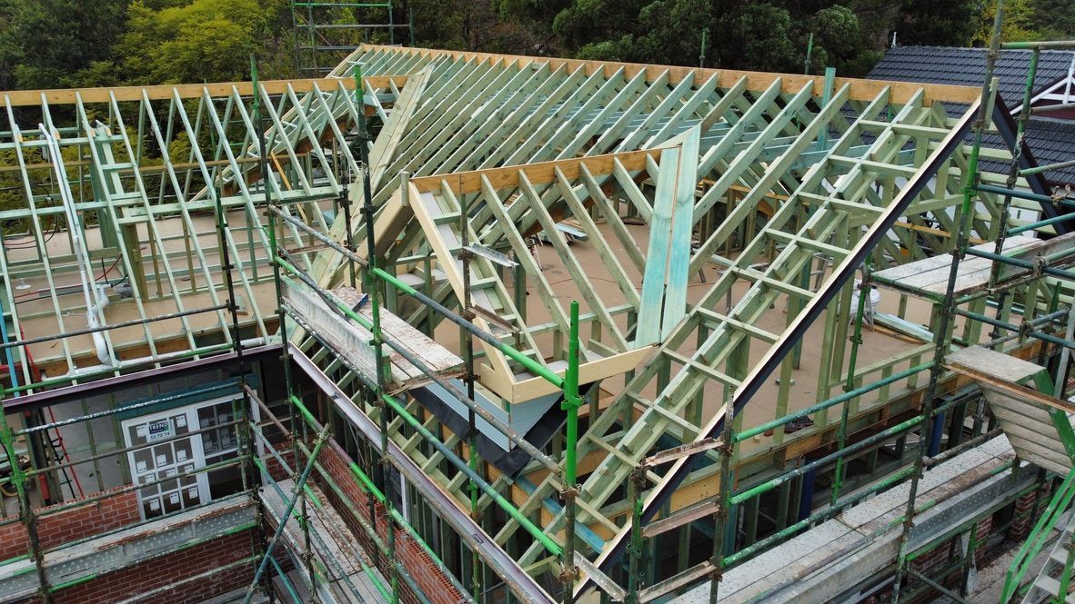 Wooden roof framing under construction around a house, with scaffolding and green safety netting.