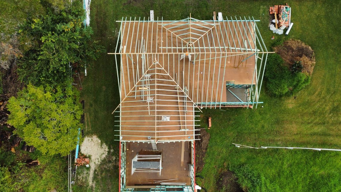 Aerial view of a house under construction with exposed roof framing and surrounding trees.