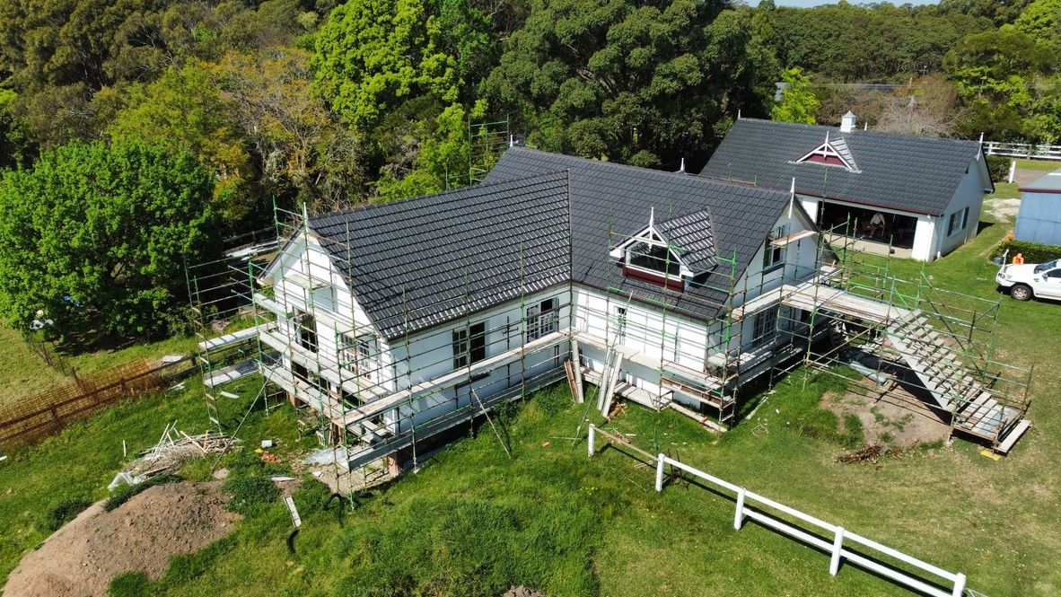 Aerial view of a white house with a dark roof and wraparound porch in a grassy wooded setting
