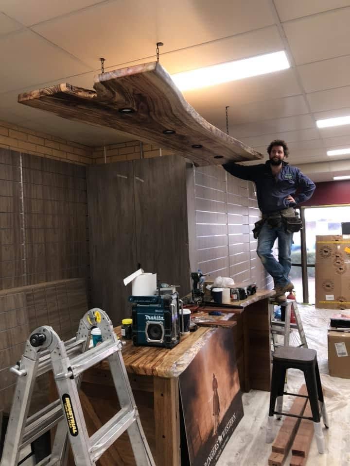 Man standing on a ladder installing a large ceiling panel in a tiled room with tools and ladders below