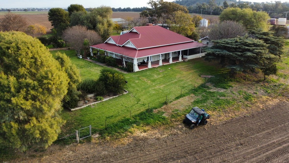 A red-roof farmhouse with a green lawn, surrounded by trees and fields, seen from above.