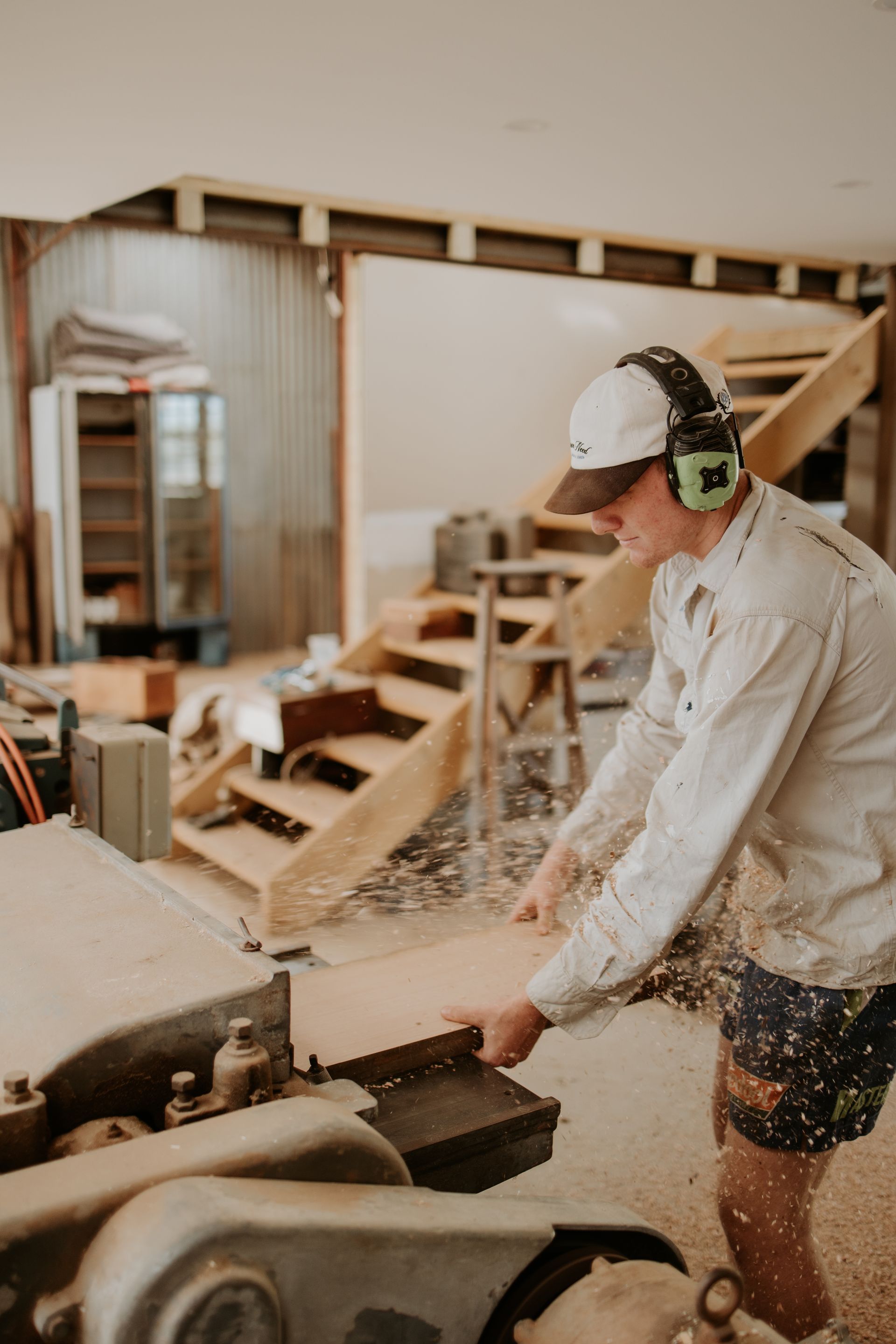 Worker cutting wood in a workshop, wearing safety earmuffs and gloves, with sawdust flying.