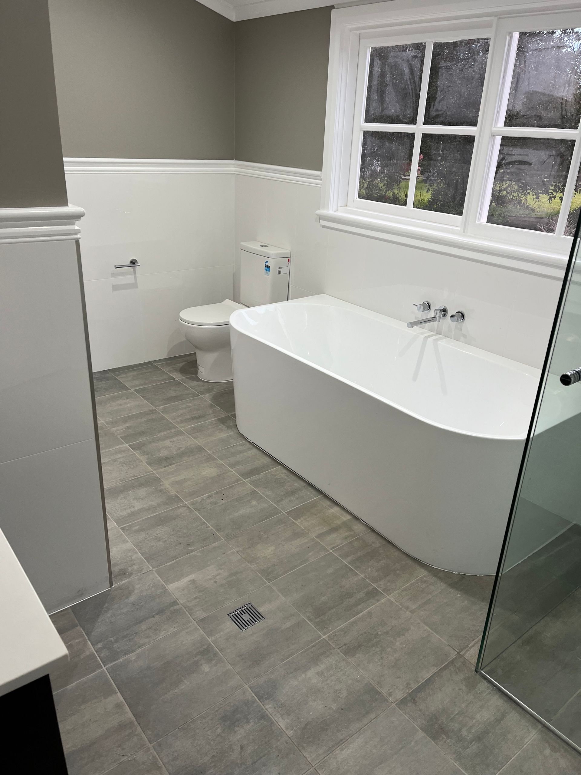 Modern Bathroom With White Tub, Toilet, Gray Tile Floor, and a Window — Boyne Building & Renovations in Ponto, NSW