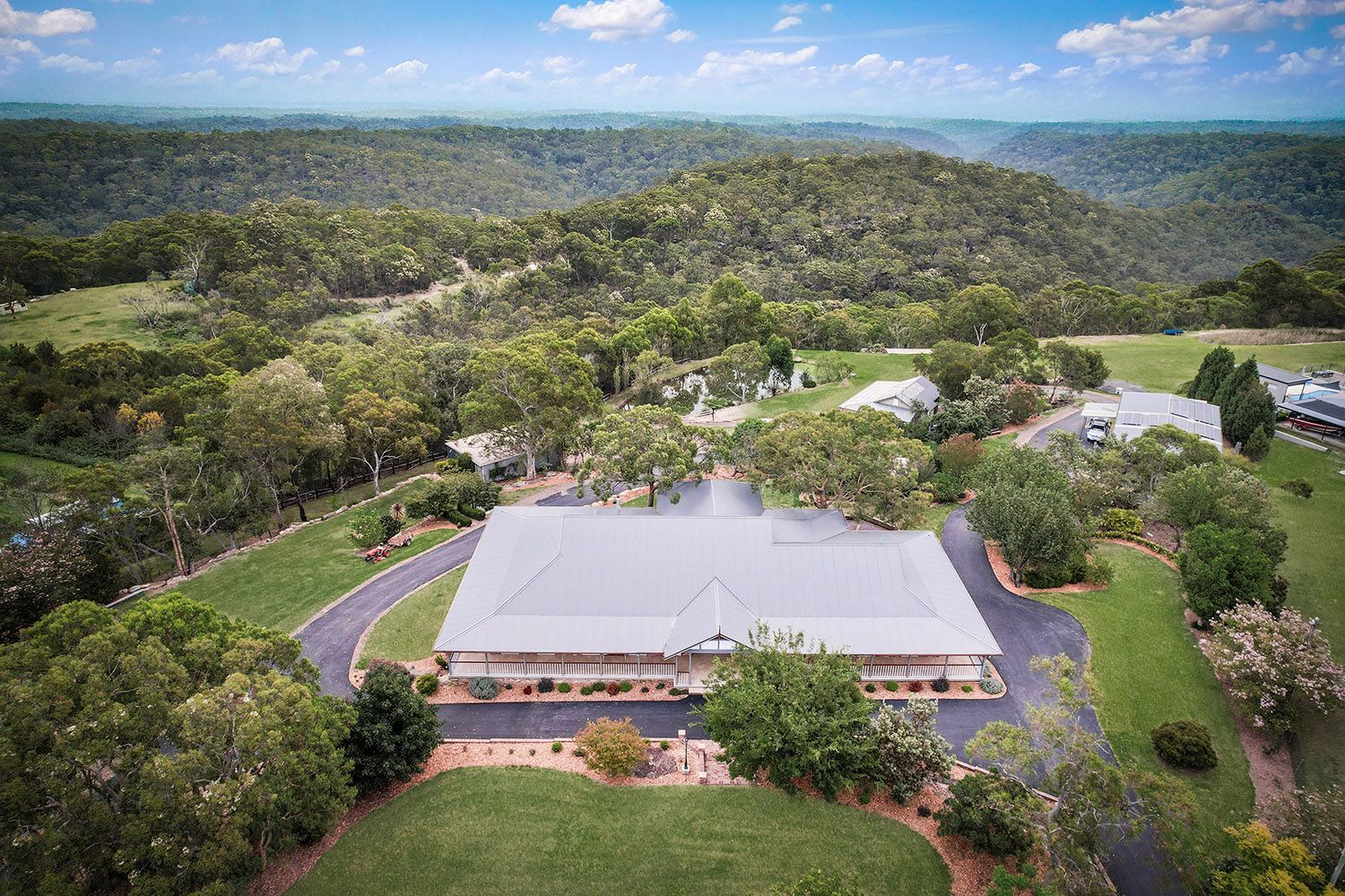 Aerial view of a large house surrounded by trees and hills under a partly cloudy sky.