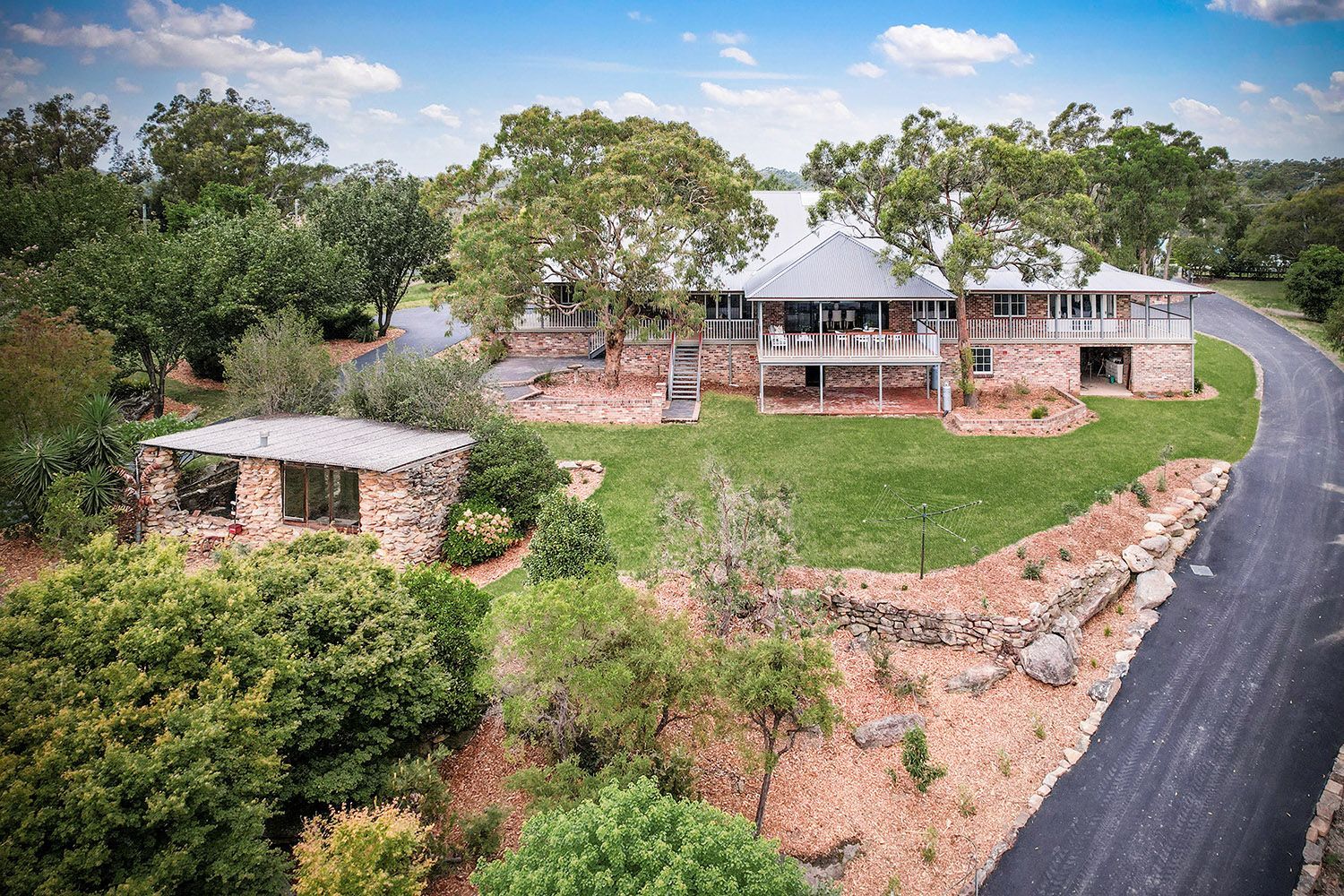 Stone house with a wraparound porch on a grassy lot beside a curved paved road, seen from above