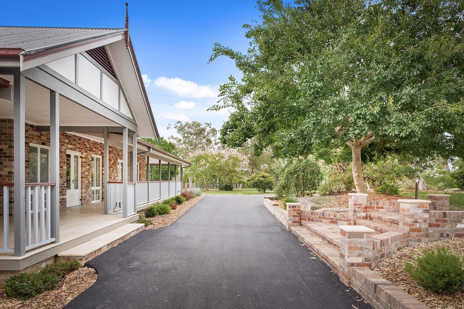 Tree-lined driveway beside a white building and stone wall under a blue sky