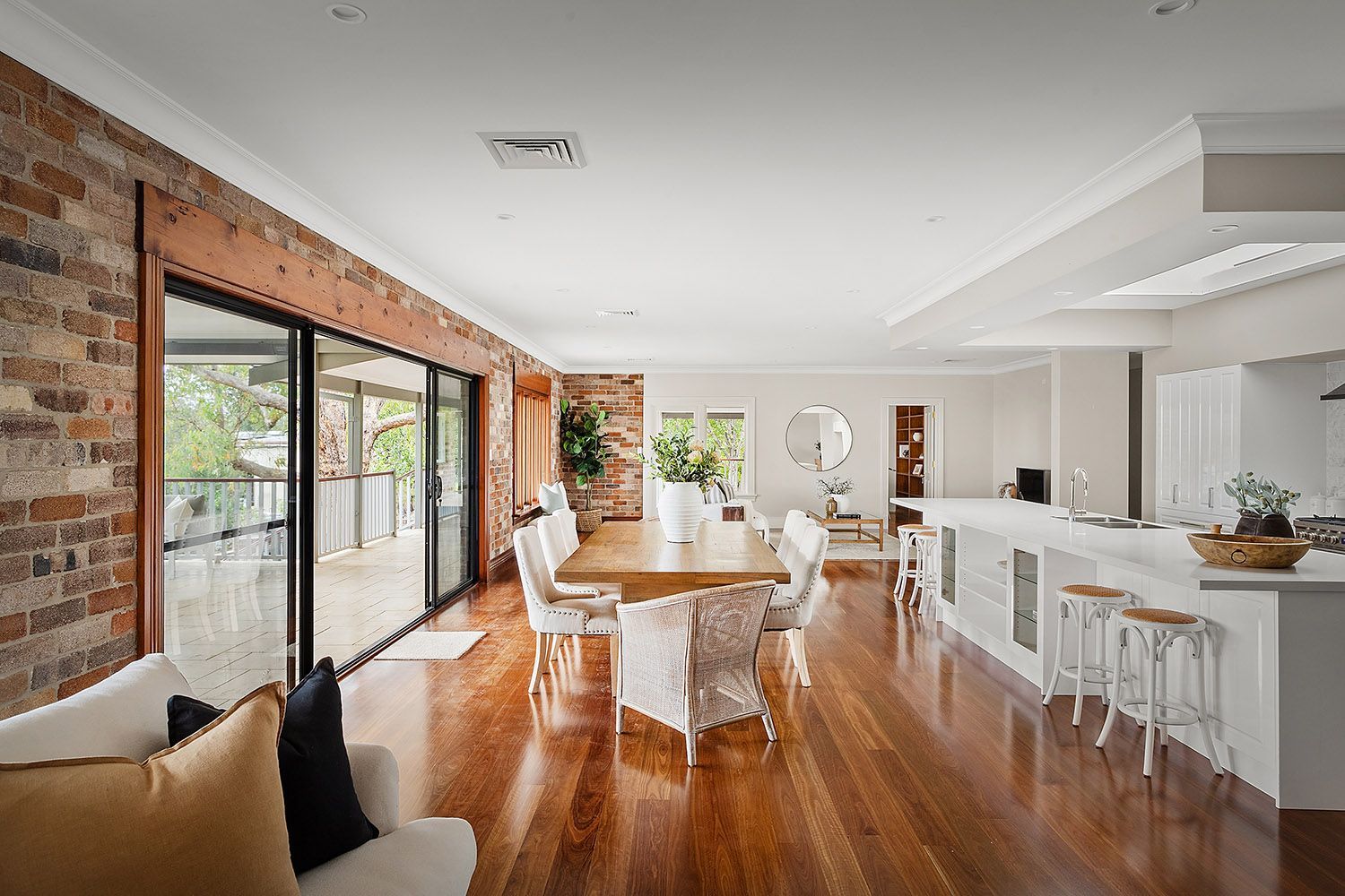 Bright open-plan living and dining room with wooden floors, brick wall, sliding glass doors, and white kitchen barstools