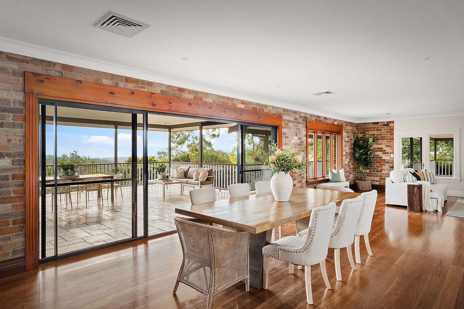 Modern dining room with brick walls, glass doors, white chairs, and a long table overlooking a patio.