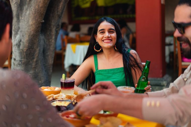 Woman in a green dress smiles at a table outdoors, holding a drink in a lively restaurant setting