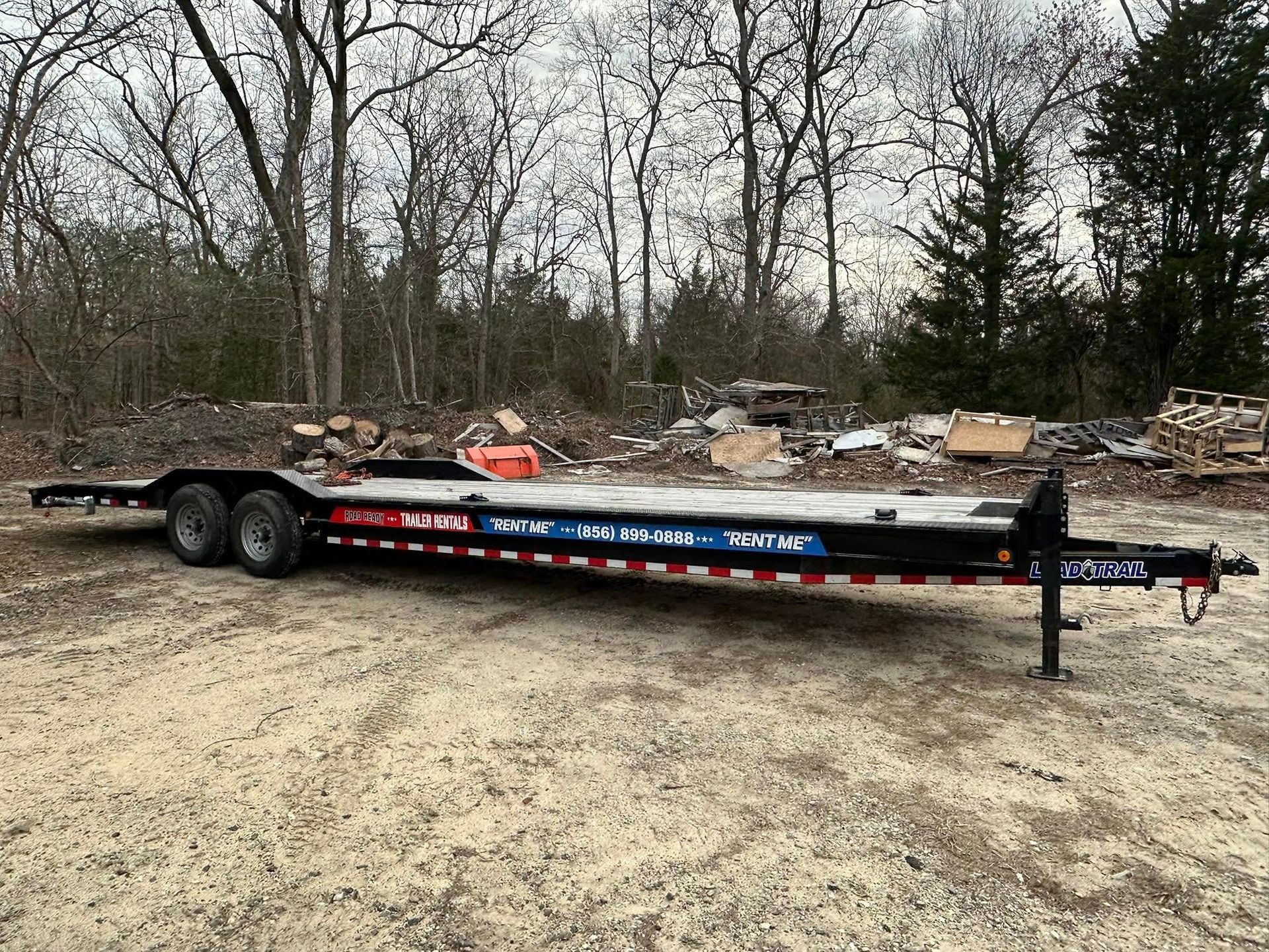 A black, dual-axle flatbed equipment trailer parked on a gravel lot in front of a wooded area.