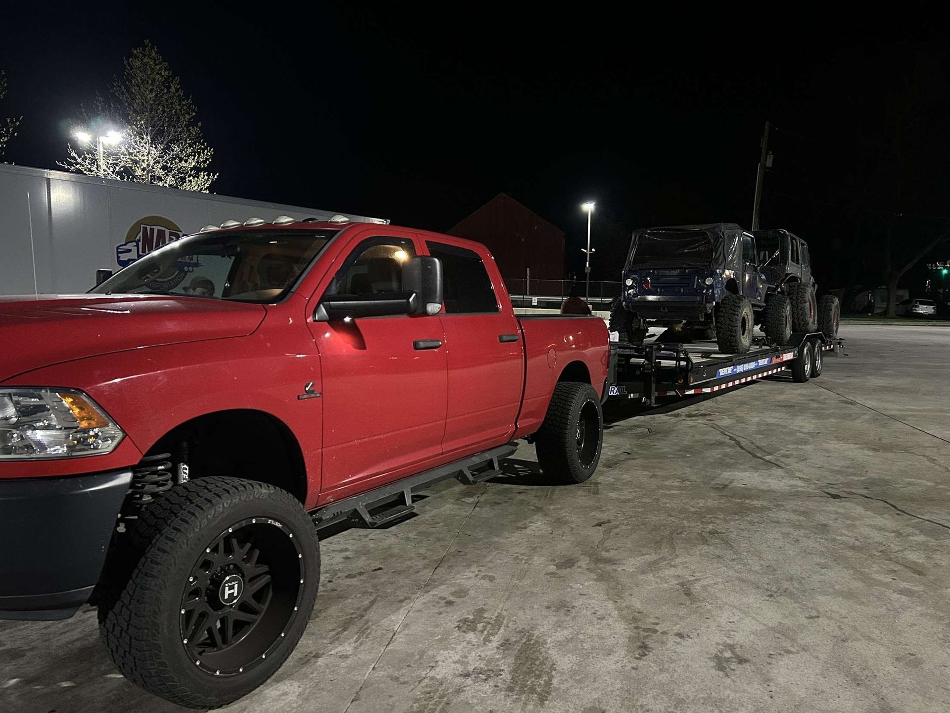 A red pickup truck is parked at night, towing a flatbed trailer loaded with two dark-colored off-road vehicles.