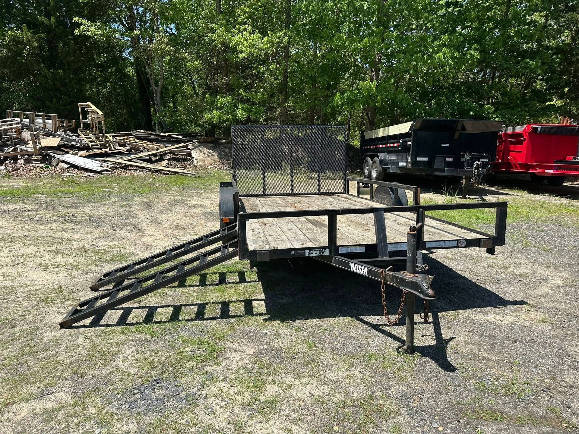 A black metal utility trailer with a wooden deck and attached loading ramps, parked on gravel in a yard.
