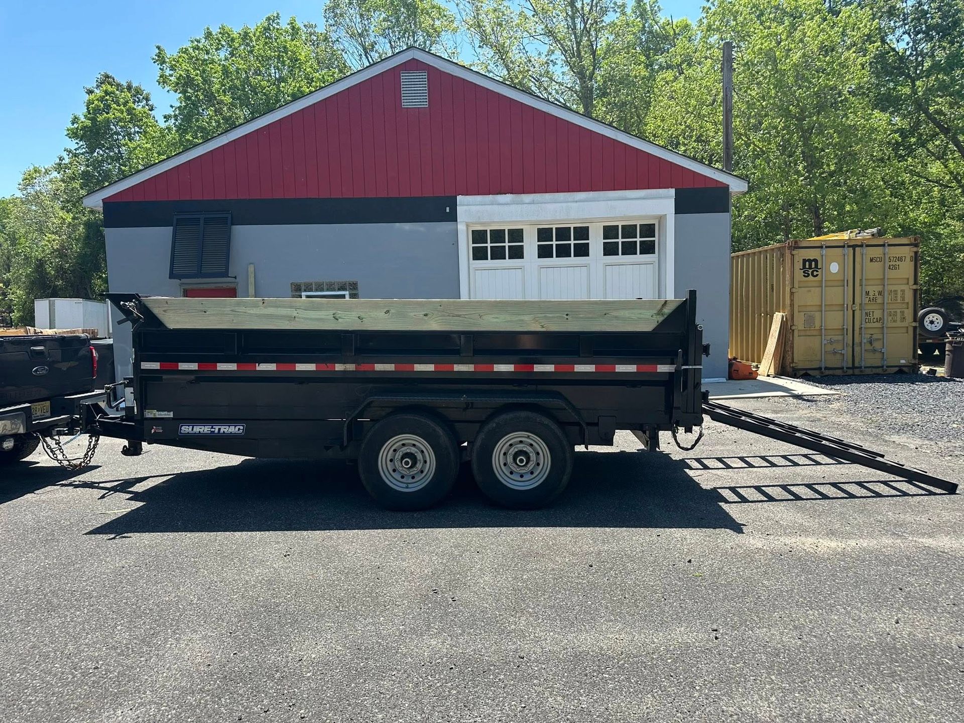 A black dump trailer parked on a gravel lot in front of a building with a red gable roof and gray walls.