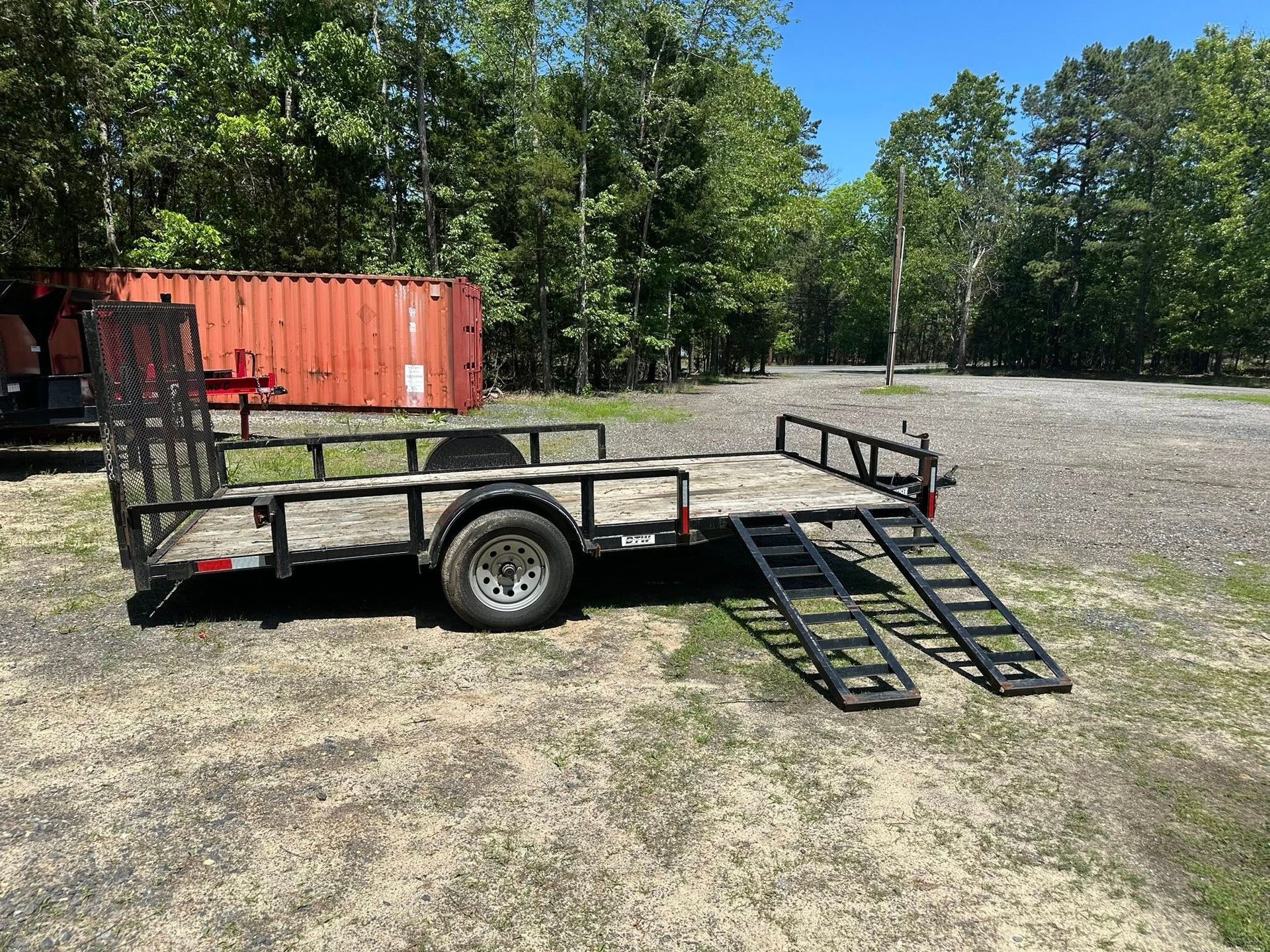 A black flatbed utility trailer with a mesh gate and two fold-down ramps sits on a dirt lot near a red shipping container.