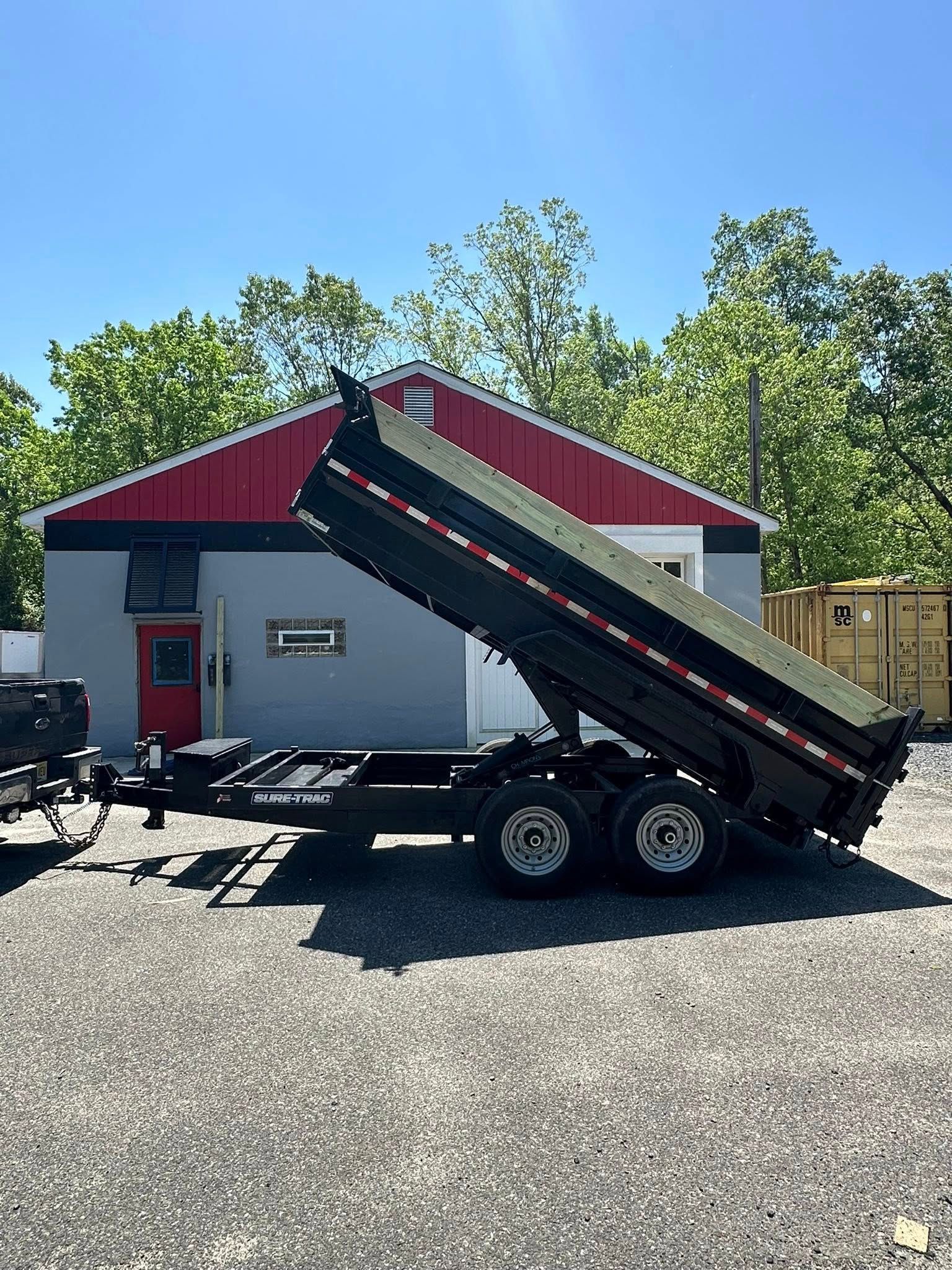 A black dump trailer in an elevated, tilted position parked on a gravel lot in front of a building with red and grey walls.